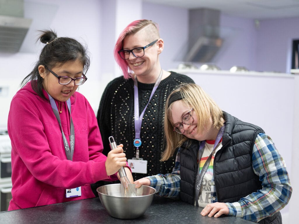 Two inclusive learners mixing a bowl of ingredients in a kitchen with a lecturer, smiling.