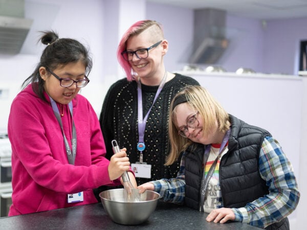 Two inclusive learners mixing a bowl of ingredients in a kitchen with a lecturer, smiling.