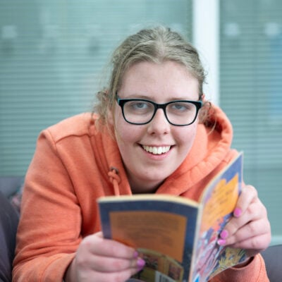 Inclusive learning student smiling at the camera whilst reading a book. She is wearing glasses and a orange hoodie.